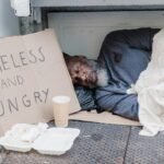 Elderly homeless man sleeping on the street with a sign reading 'Homeless and Hungry'.